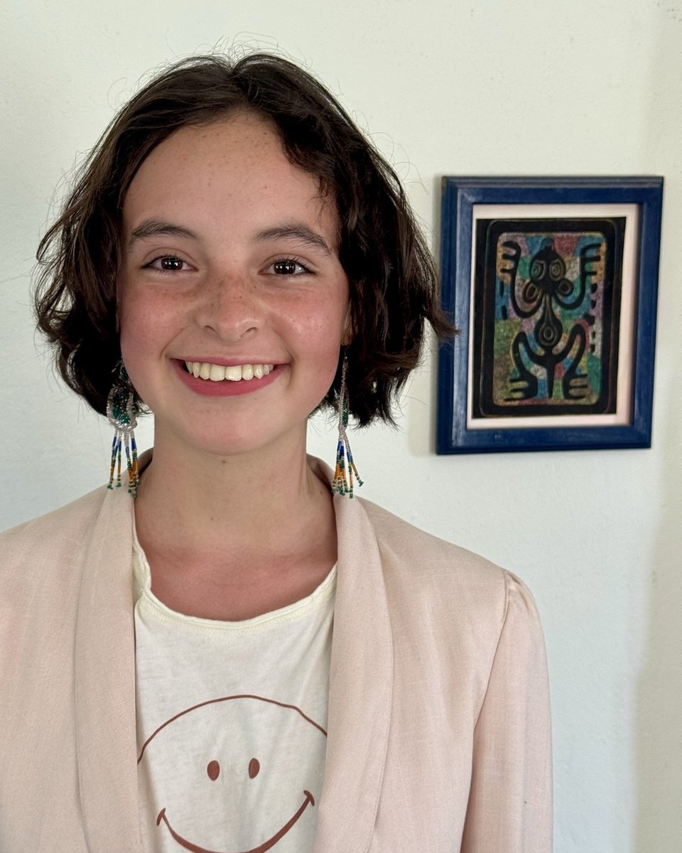 Zuri sonriendo, con saco rosa claro, frente a una pared blanca.